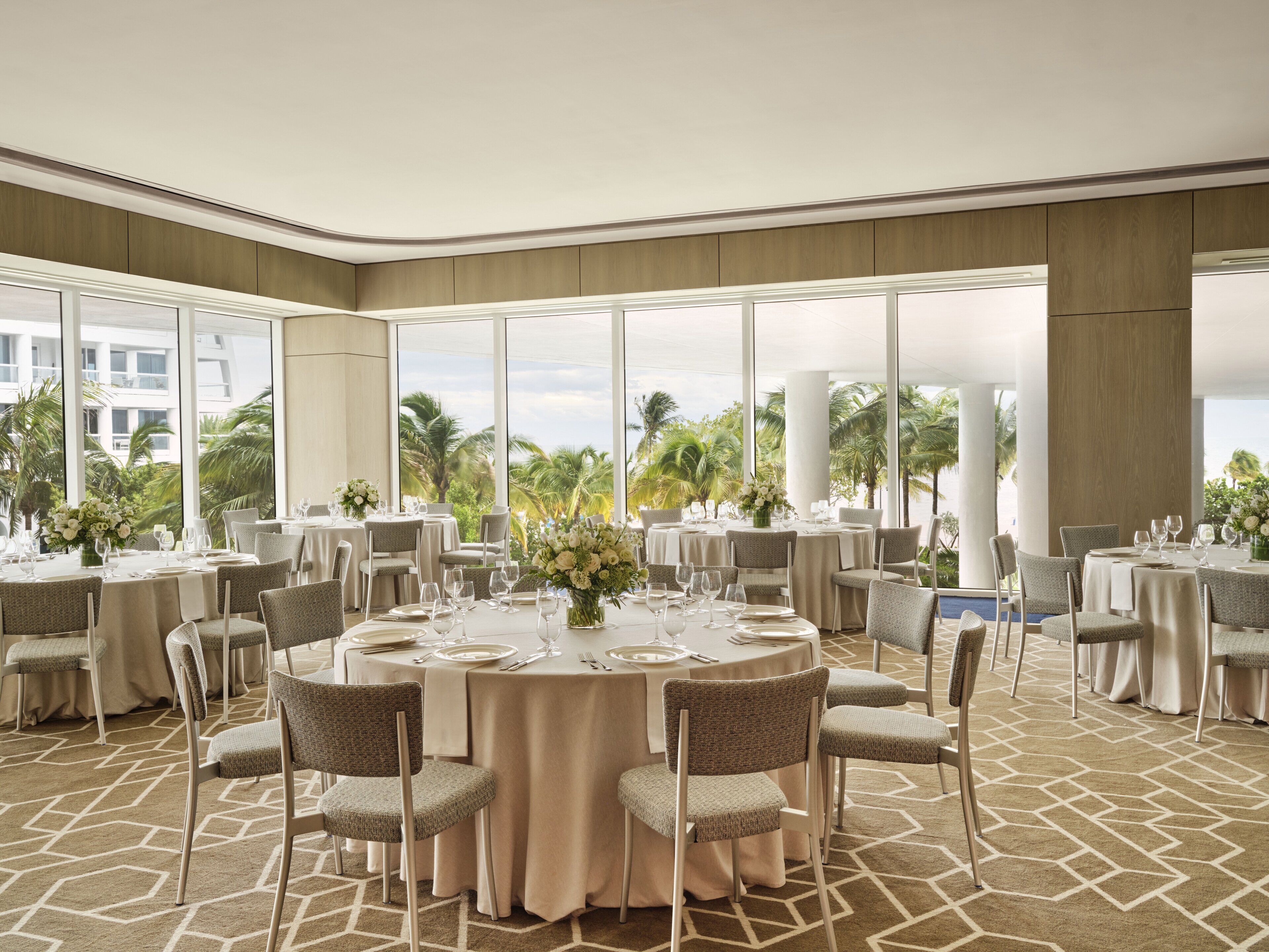 Un amplio salón de banquetes con mesas redondas y sillas, grandes ventanales que ofrecen vista a palmeras y un edificio.