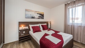A neatly made bed with a red quilt, a bedside table with a lamp, and a painting on the wall. - Pool Apartments Banta B (Novalja)