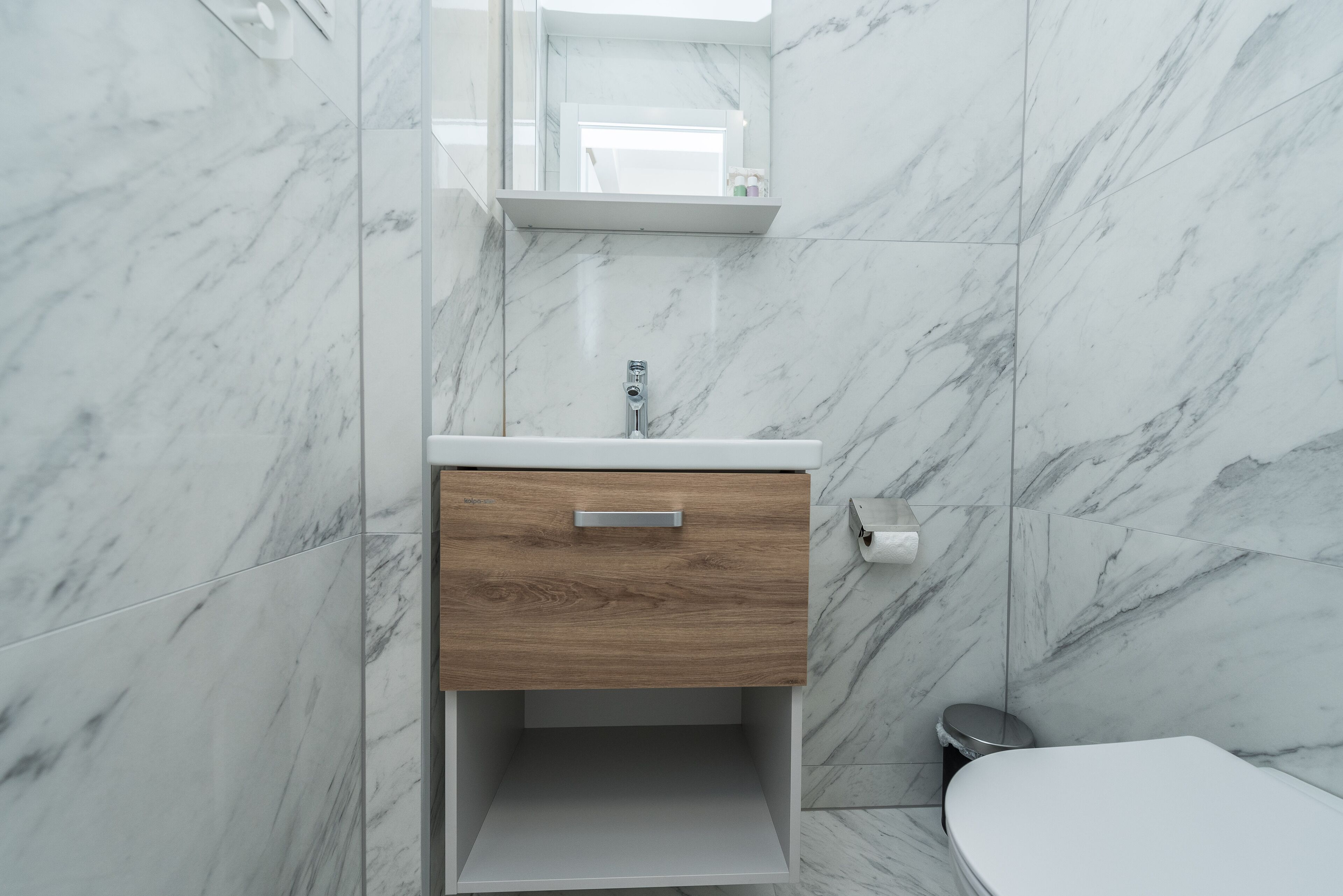 A bathroom with a marble wall, a wooden vanity with a drawer, a toilet, and a mirror above the vanity.