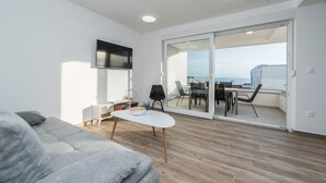 A modern living room with a sofa, a coffee table, and a TV on the wall. There is a sliding glass door leading to an outdoor balcony with a dining table and chairs. - Pool Apartments Banta B (Novalja)
