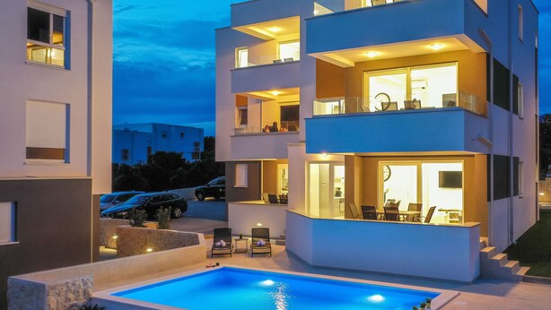 A modern multi-story building with a swimming pool, outdoor seating, and a well-lit interior. - Pool Apartments Banta B (Novalja)