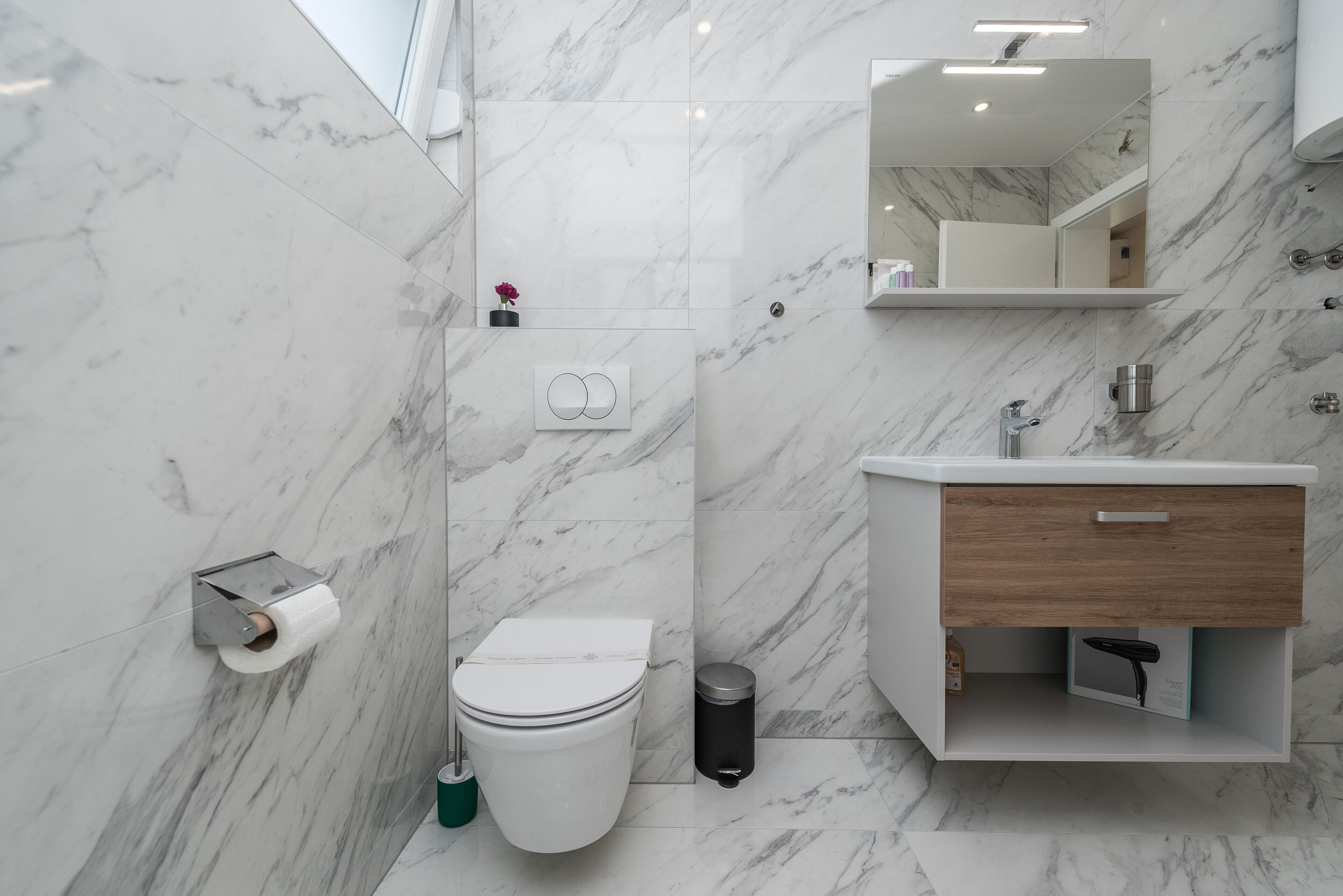 Une salle de bain moderne avec un sol en marbre, des toilettes, un lavabo avec un meuble en bois, un miroir et un porte-rouleau de papier toilette fixé au mur.