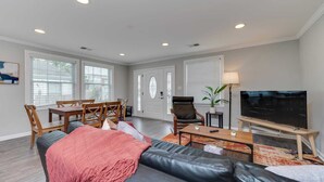 Ein modernes Wohnzimmer mit einer schwarzen Ledersofa, einem Holzesstisch und Stühlen. Ein Fernseher steht auf einem Holzgestell und eine Stehlampe ist vorhanden.