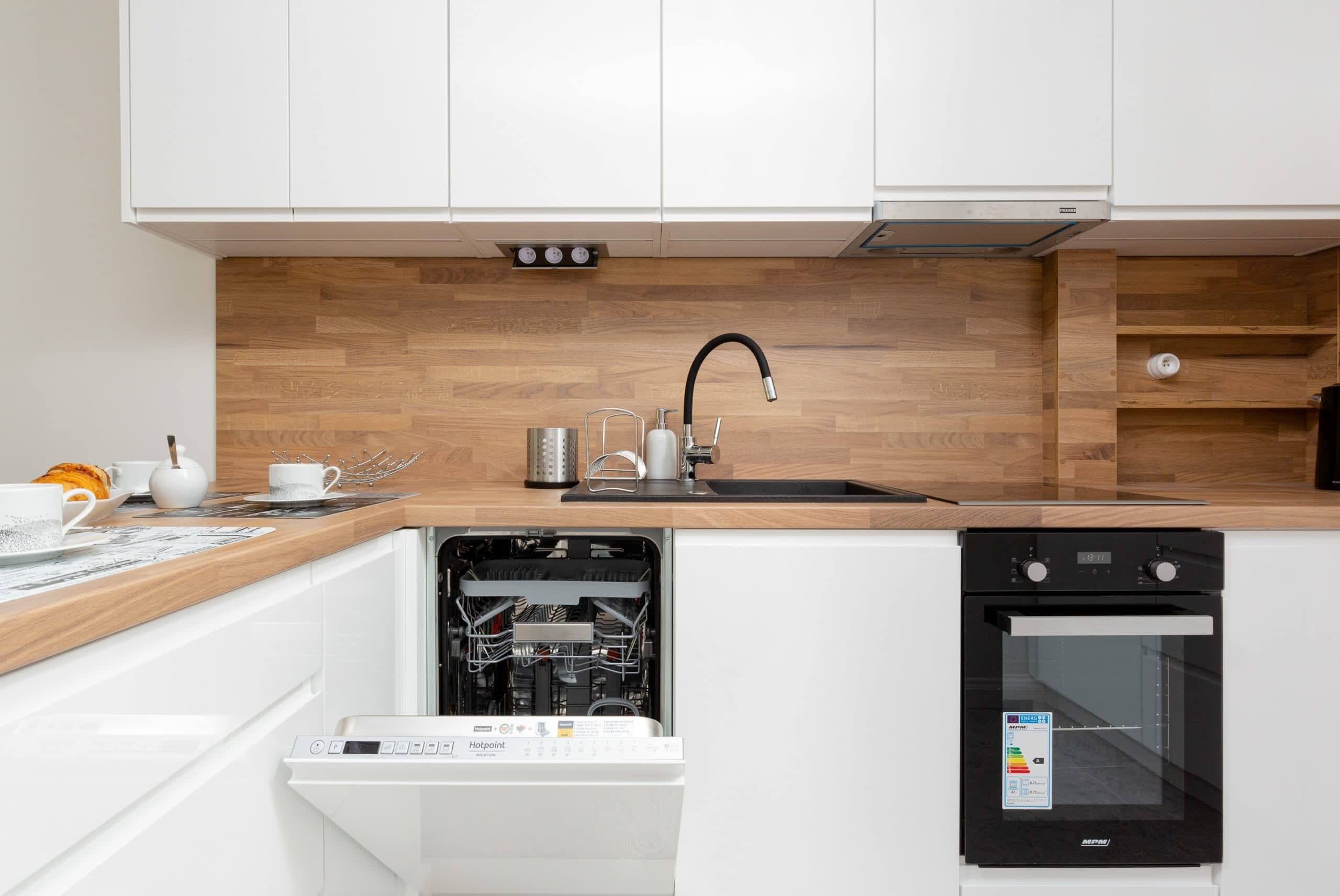 Cocina compacta con armarios blancos, protector contra salpicaduras de madera, lavavajillas integrado y horno empotrado.