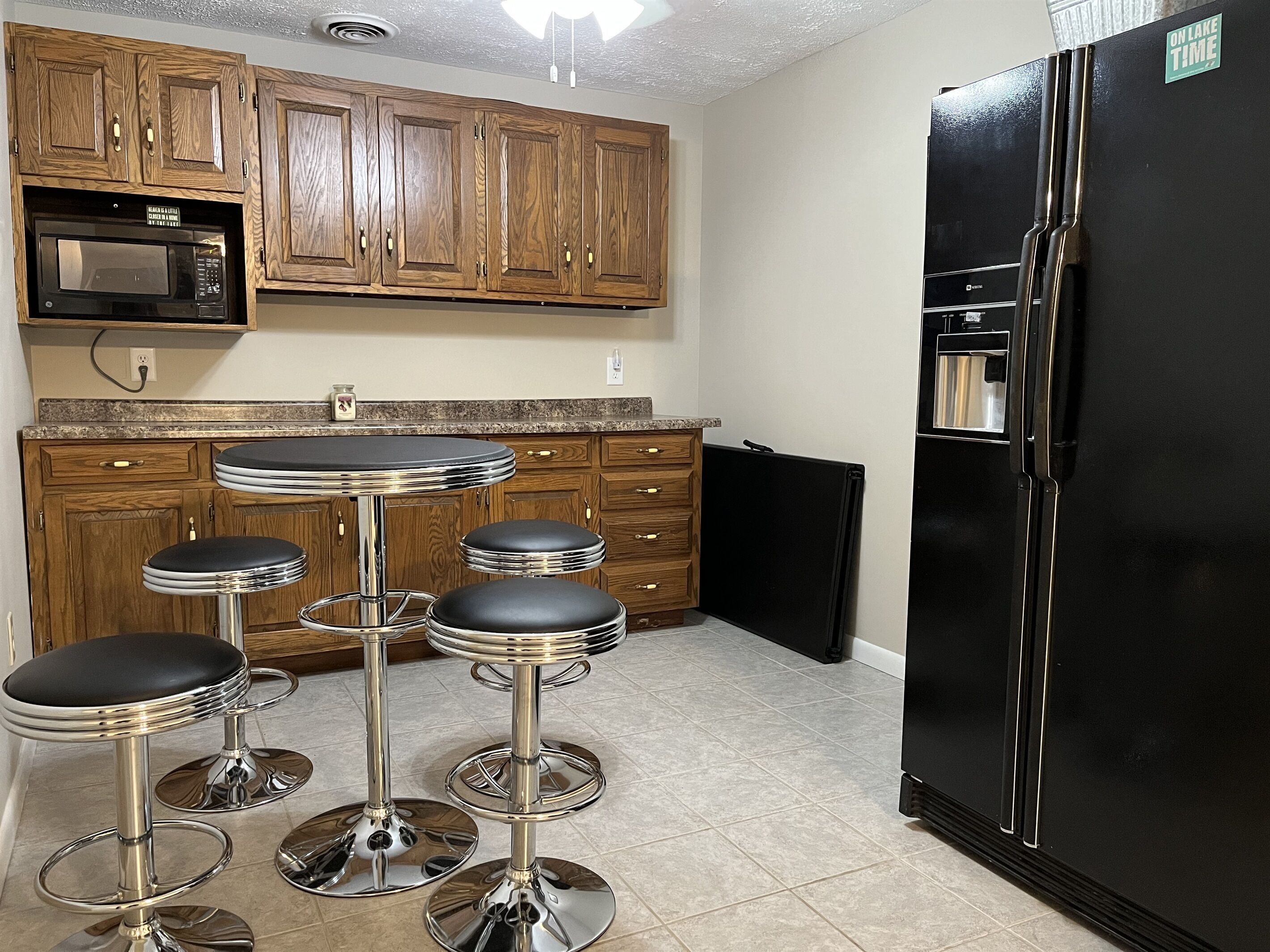 A kitchen with wooden cabinets, a black refrigerator, a microwave, and three bar stools.