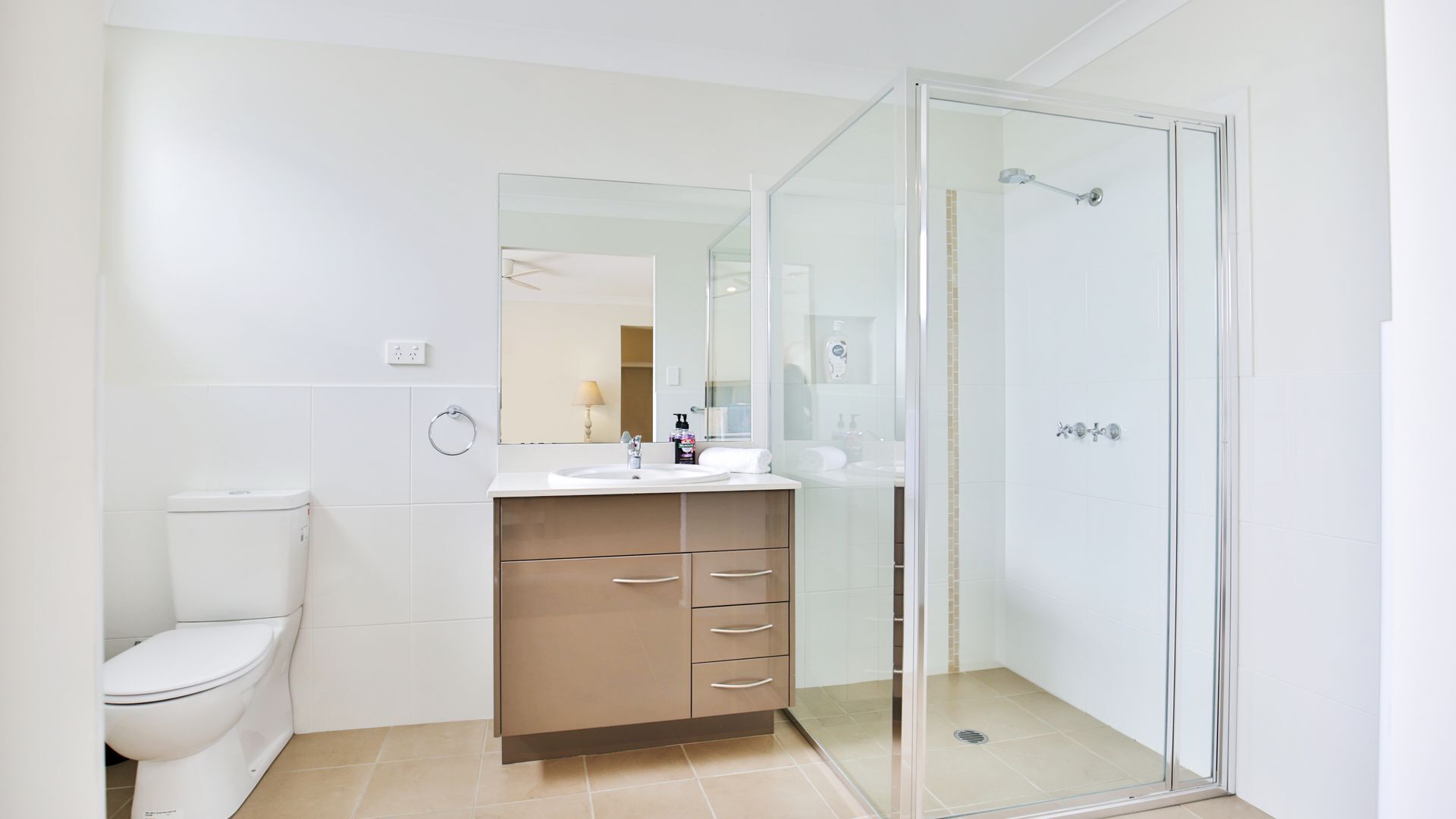 Un baño moderno con inodoro, lavamanos con espejo, cabina de ducha de cristal y un mueble beige con cajones.