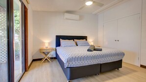 Une chambre à coucher comprenant un lit, des tables de chevet, un appareil de climatisation et une vue sur l’extérieur, visible à travers une porte vitrée.