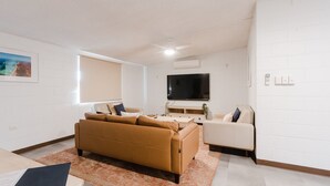 Un salon moderne avec un canapé marron, un fauteuil blanc, un téléviseur à écran plat et une grande fenêtre équipée de stores.