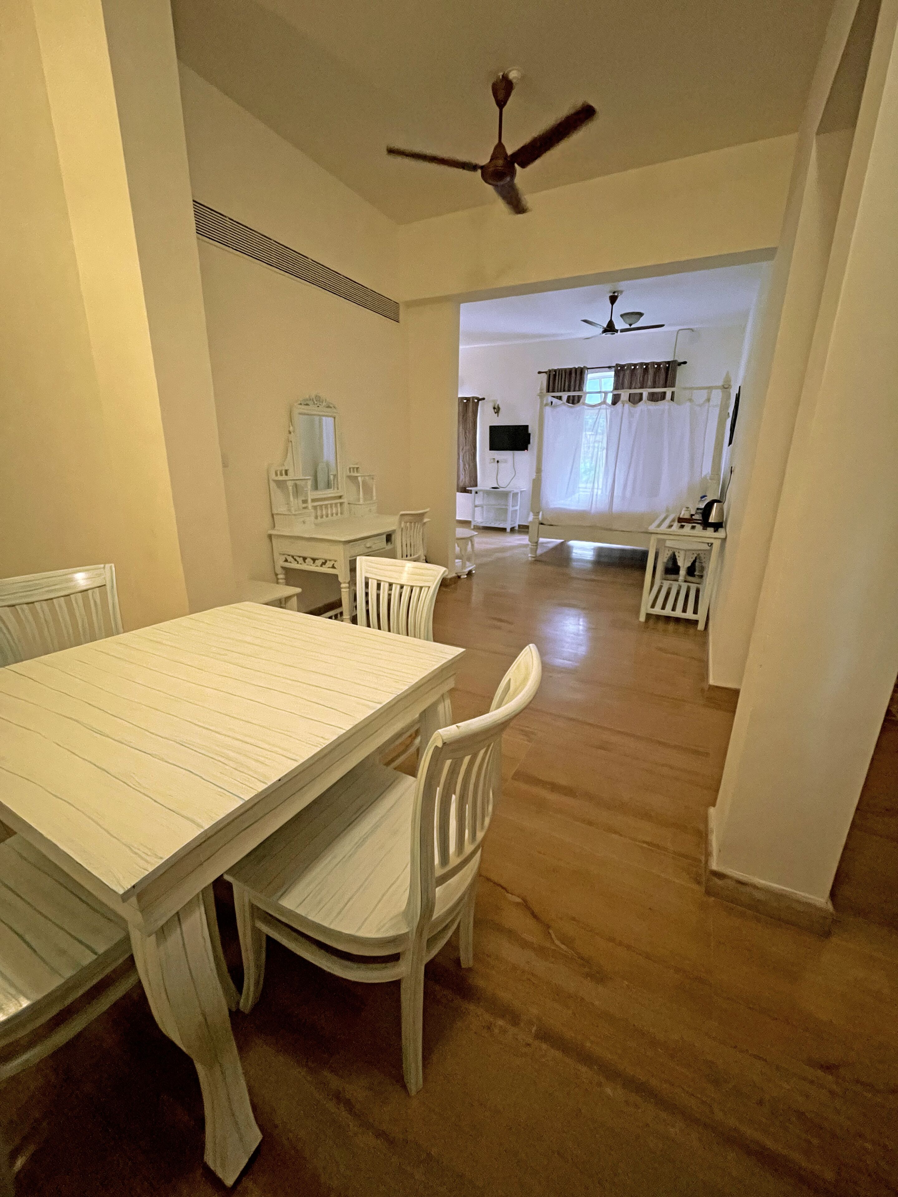 A dining area with a white table and chairs, a ceiling fan, and a view into a bedroom with a bed and curtains.