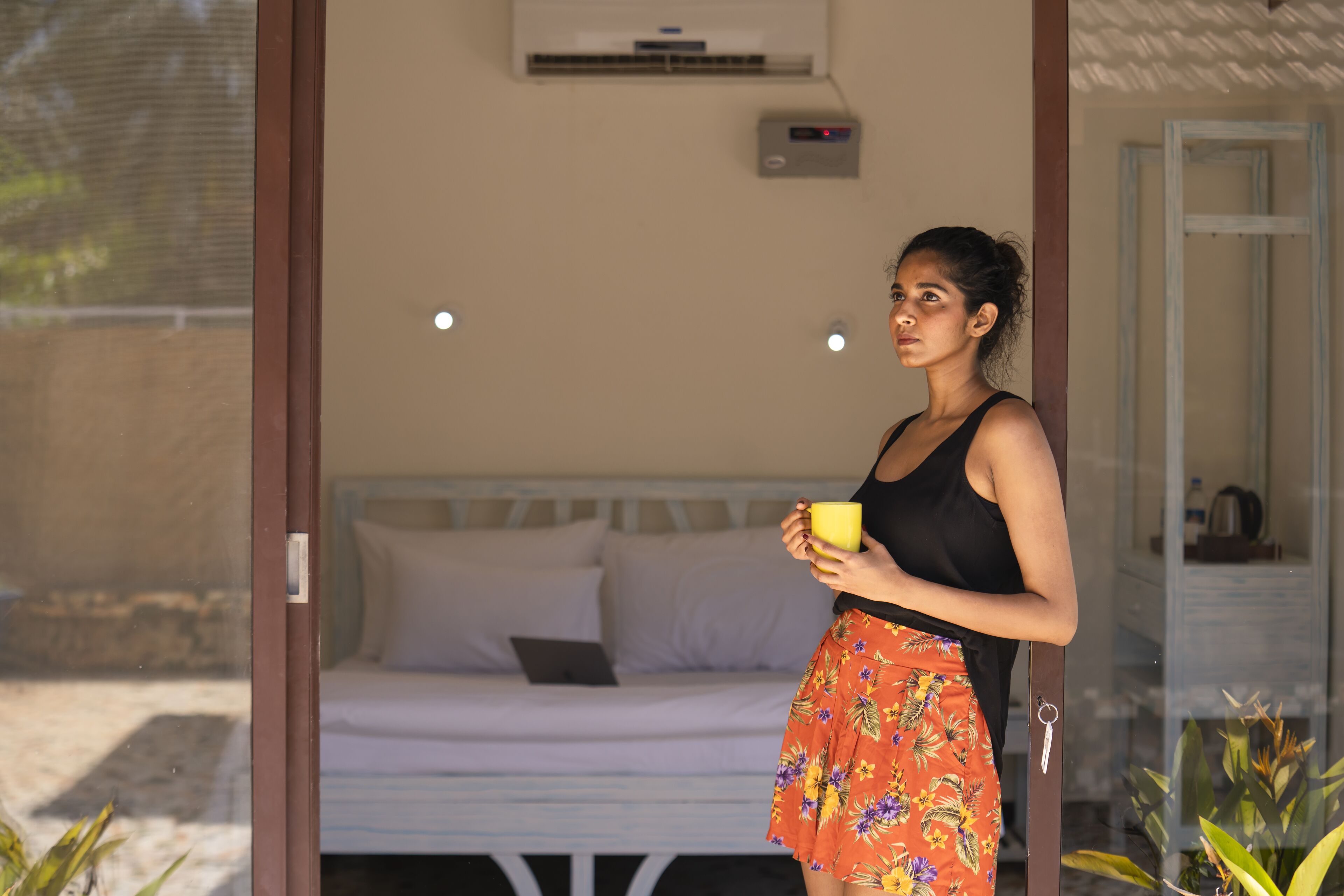 A person standing in a doorway holding a yellow mug, with a bed and a small table visible in the background.
