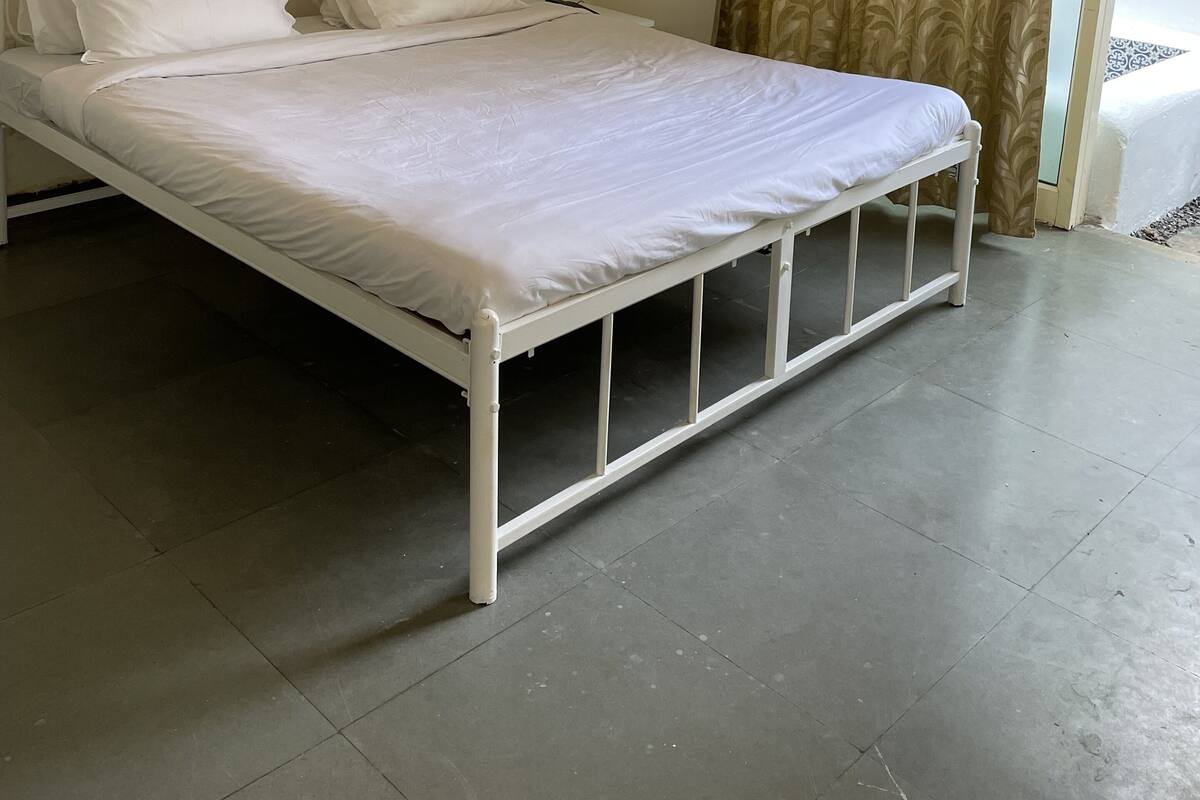 A single-bed room with a white metal frame, a ceiling-mounted air conditioner, a framed picture on the wall, and a patterned curtain on the window.