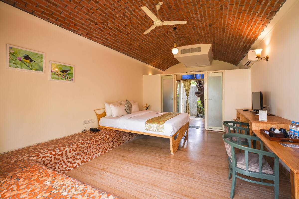 A bedroom with a bed, a desk, chairs, a ceiling fan, and a brick ceiling.