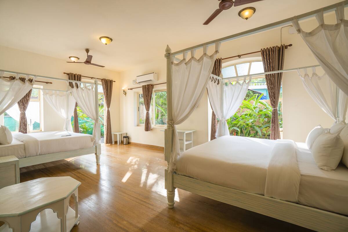 A four-poster bed with white bedding and curtains, a ceiling fan, and a view of greenery outside.