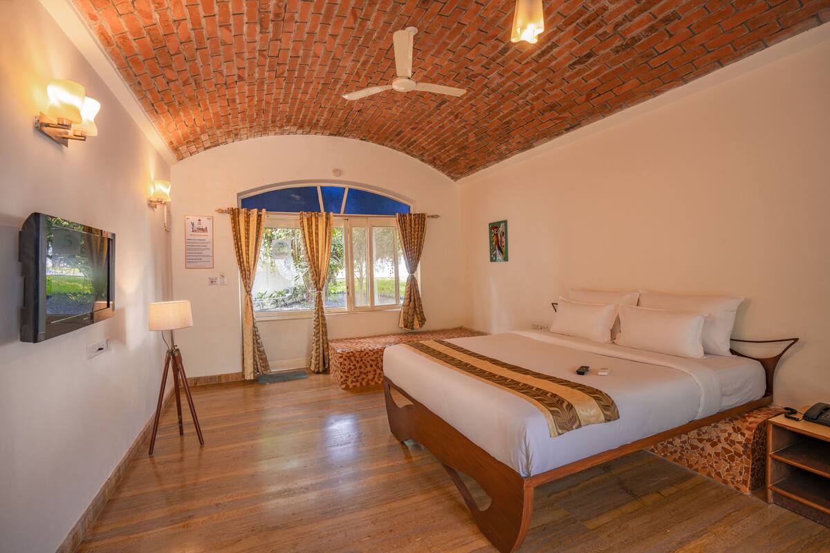 A room with a brick ceiling, a bed with white bedding, a wooden floor, a standing lamp, and a television on the wall.
