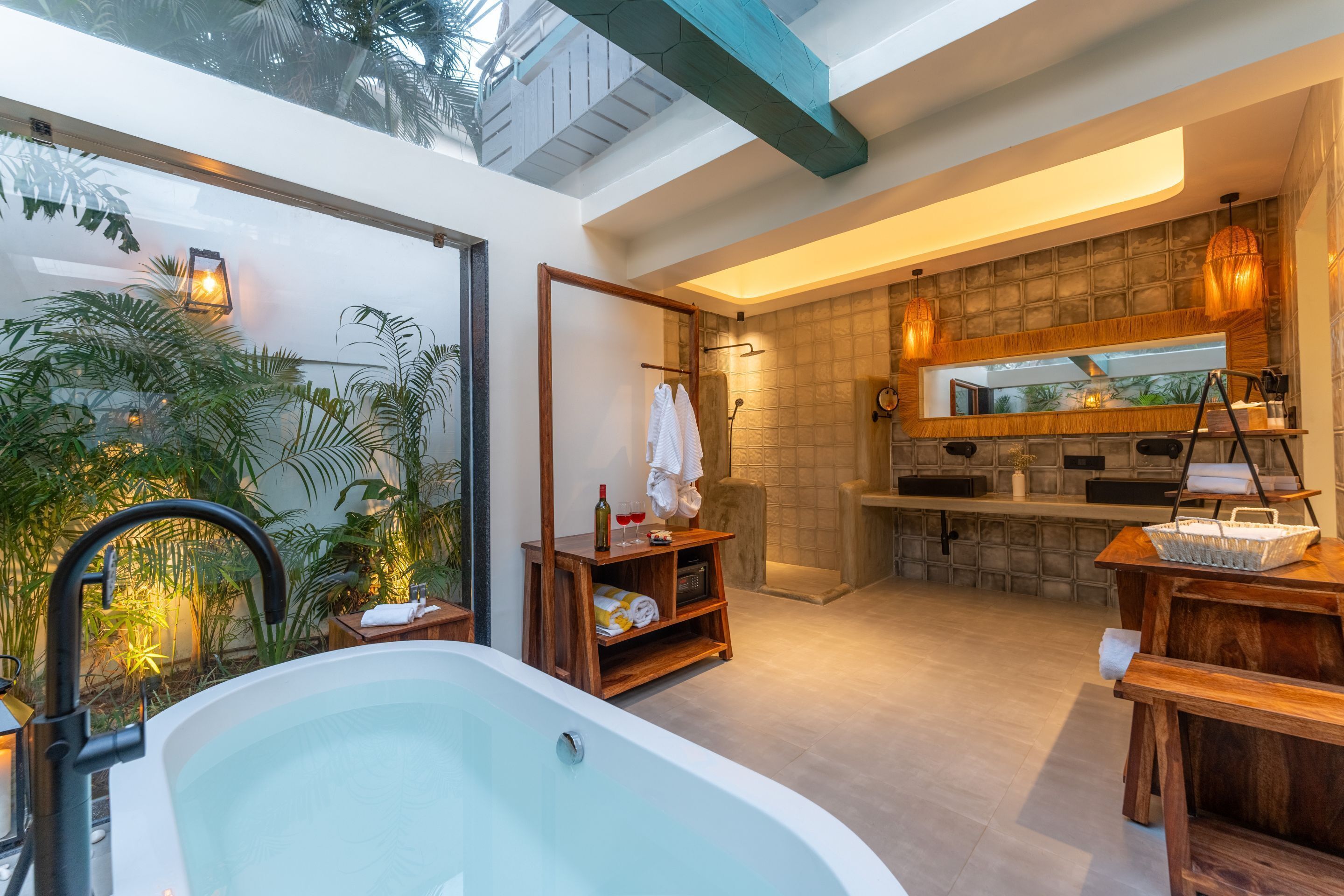 Un baño moderno con una bañera grande, zona de ducha y tocador con espejo. Hay plantas y un estante con toallas y artículos de aseo.