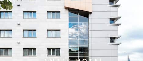 Een modern hotel met een opvallende glazen gevel, meerdere balkons en een centrale ingang met terraszitplaatsen.