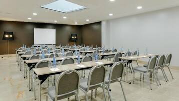 Un salón de conferencias con varias mesas y sillas, equipado con botellas de agua y documentos, una pantalla de proyector y una lámpara en el techo.