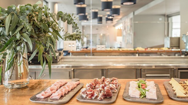 A buffet with various meats and cheeses, a vase with green leaves, and a modern kitchen in the background. - Hotel Antik San Sebastian (San Sebastián)