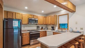A modern kitchen with wooden cabinets, stainless steel appliances, a central island with stools, and a window with a view. - Experience slope side on Salamander at The Haven! (Canaan Valley)