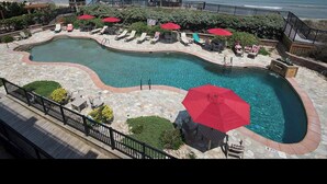 A swimming pool with a curved design, surrounded by lounge chairs and umbrellas, with a view of the ocean. - Beachside Breeze - Pools, hot tub, & private beach access (Kill Devil Hills)