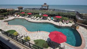 A swimming pool with a curved design, surrounded by lounge chairs and umbrellas, with a view of the ocean. - Beachside Breeze - Pools, hot tub, & private beach access (Kill Devil Hills)
