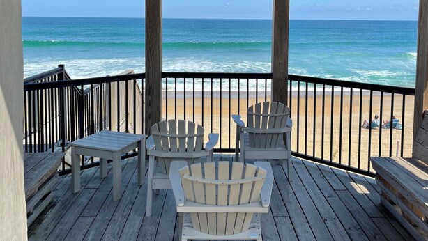 A wooden deck with Adirondack chairs and a table, overlooking a beach and ocean. - Beachside Breeze - Pools, hot tub, & private beach access (Kill Devil Hills)