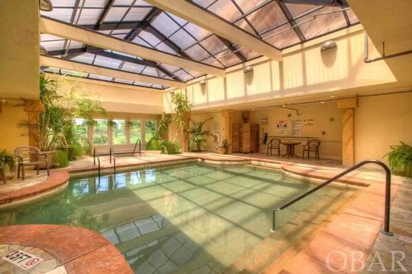 An indoor swimming pool with a glass ceiling, surrounded by plants and furnished seating areas.