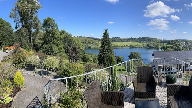 A balcony with wicker furniture overlooking a lake, surrounded by greenery and a residential area. - Dream house on the Maar (Schalkenmehren)