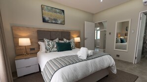 A modern bedroom with a large bed, two bedside tables with lamps, a headboard, and a painting on the wall. - Caribbean Estates - 35 Montego Bay (Port Edward)