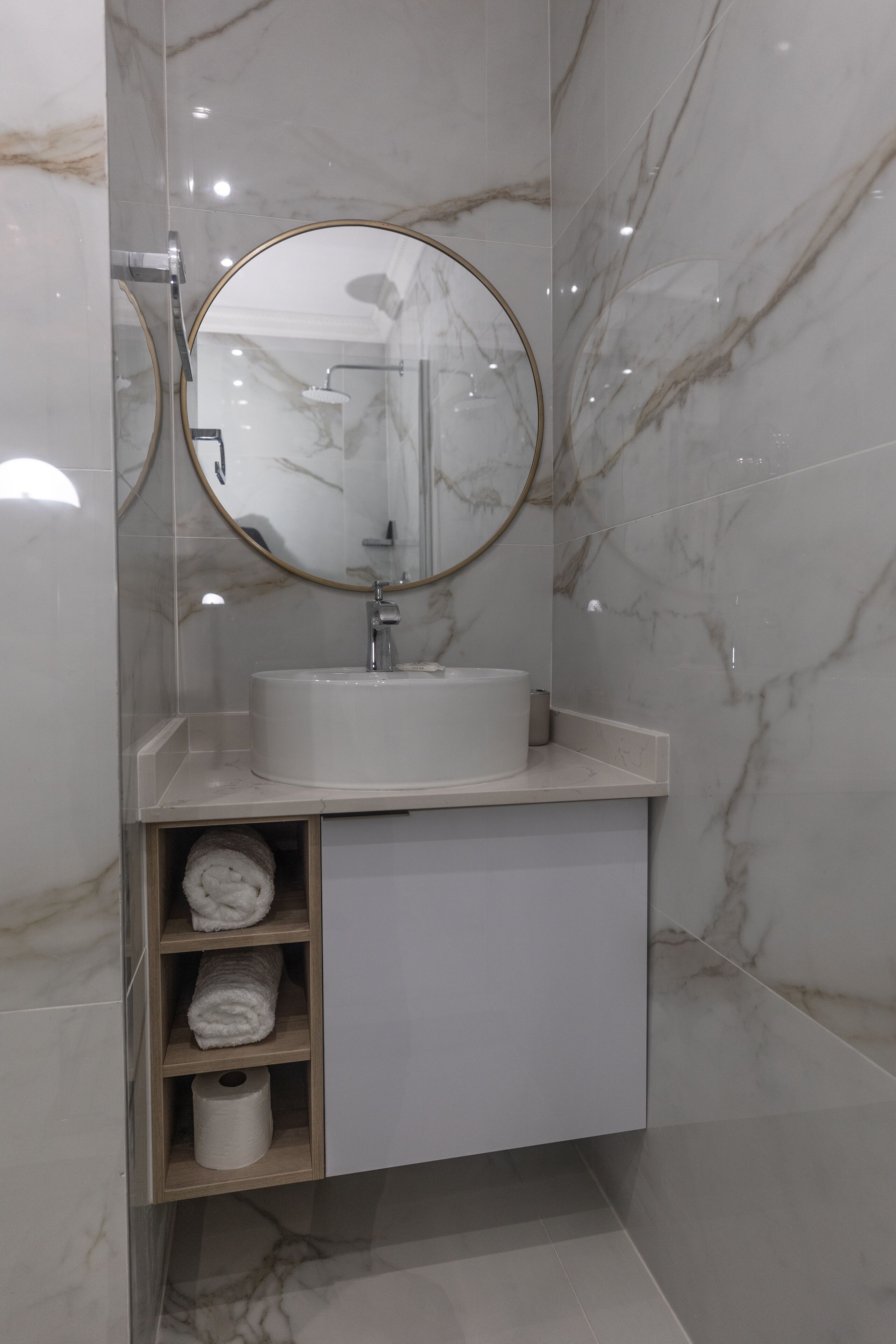 A modern bathroom with a marble wall, a round mirror, a sink, and a towel shelf.
