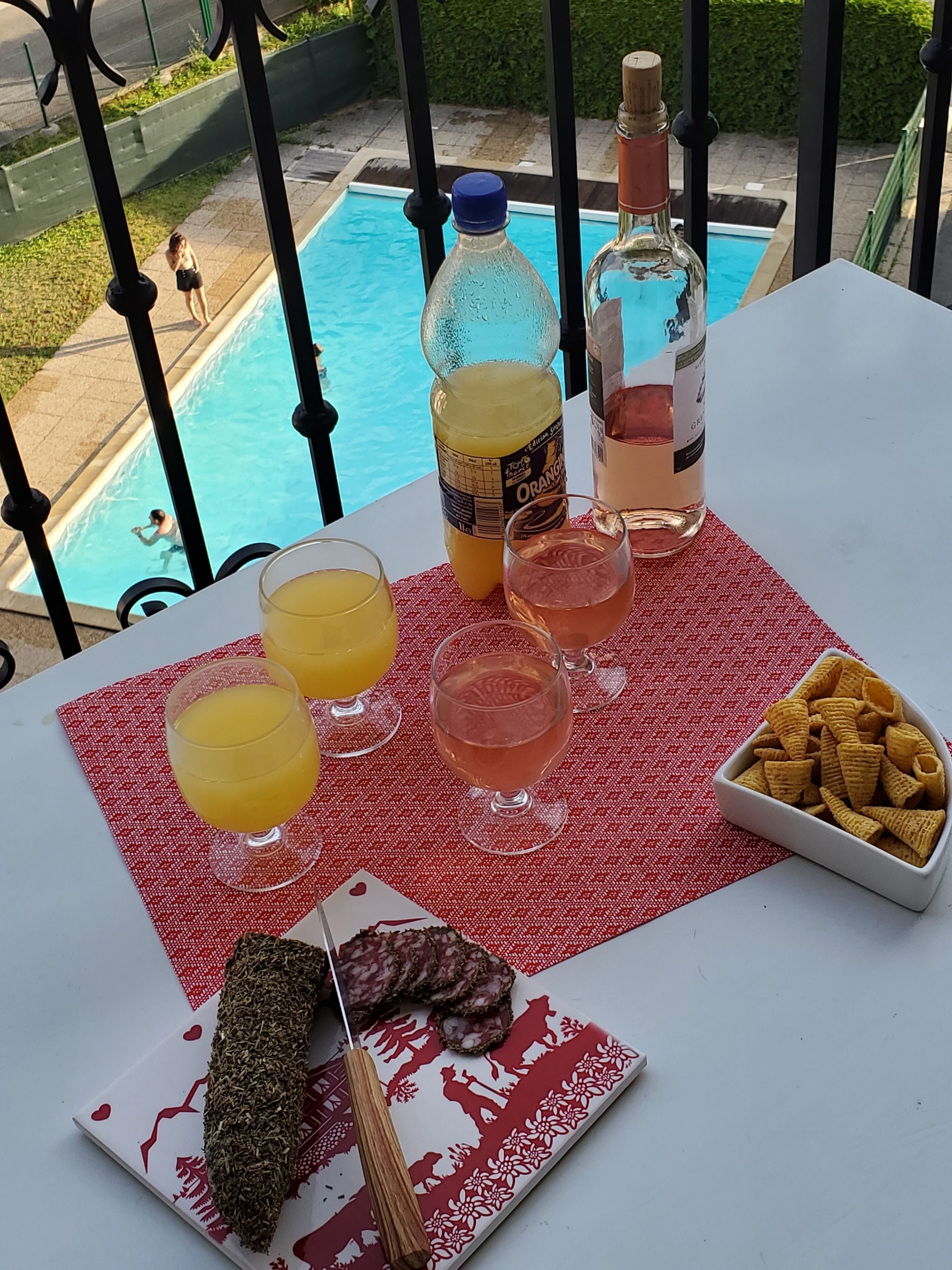 A table with drinks, snacks, and a bottle of alcohol on a balcony overlooking a swimming pool.