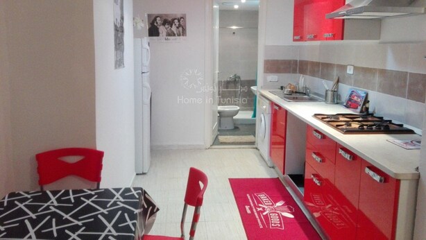 Una cocina compacta con armarios rojos, una campana extractora de acero inoxidable y una pequeña encimera. Un tapete rojo con un logotipo está en el suelo, y se ve un refrigerador blanco.