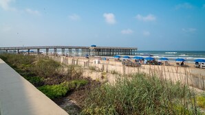 A beach with a wooden pier extending into the ocean, surrounded by beach chairs and umbrellas. - Parrots Paradise  (South Padre Island)
