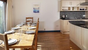 A dining area with a wooden table set for six, a kitchen with light wooden cabinets, and a view of the outdoors through a window.