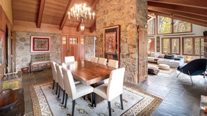 Un comedor amplio con una gran mesa de madera y sillas blancas, rodeado de paredes de piedra y vigas de madera.