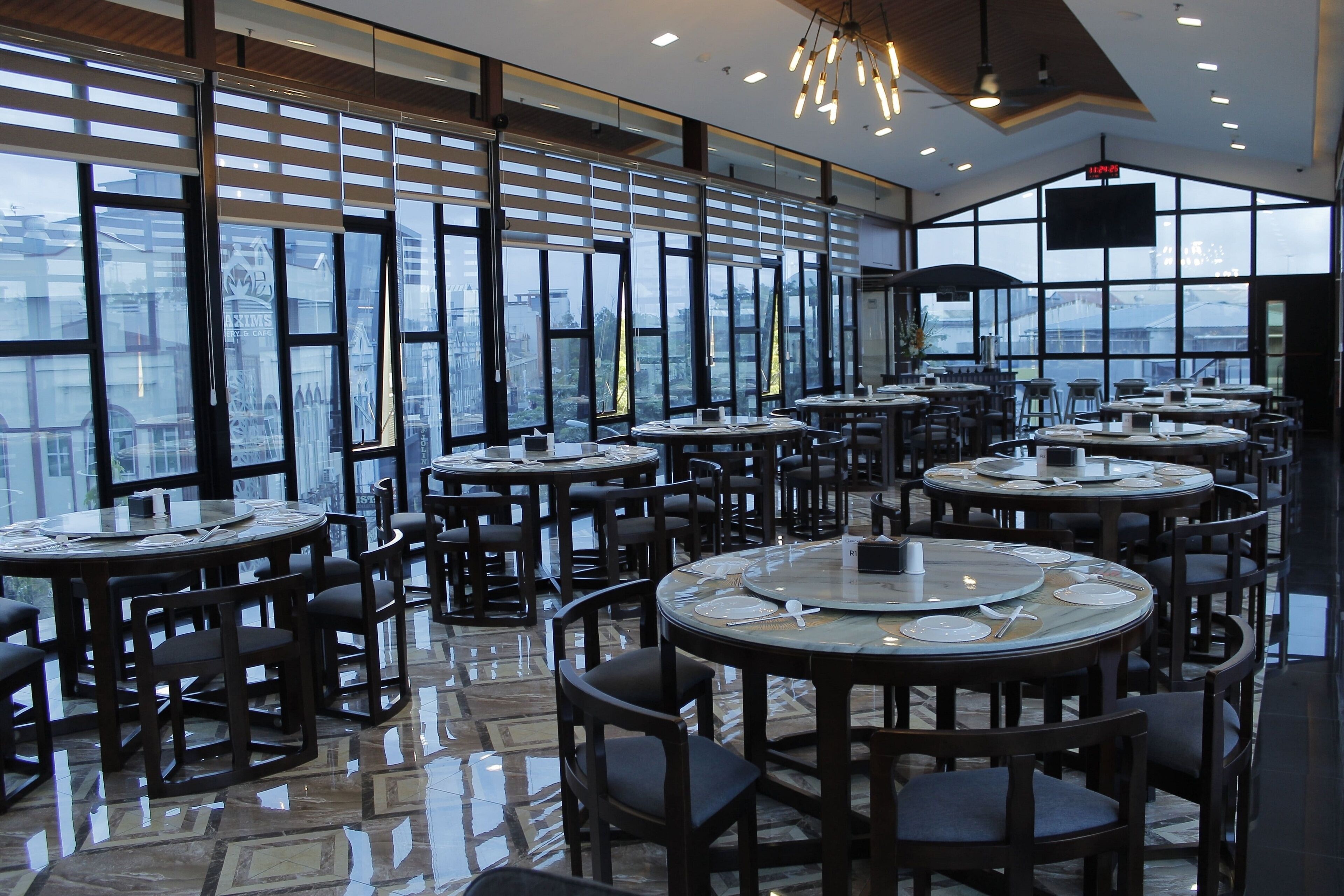 A spacious restaurant with multiple round tables and chairs, large windows offering a city view, and a modern interior design.