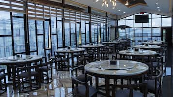 A spacious restaurant with multiple round tables and chairs, large windows offering a city view, and a modern interior design.