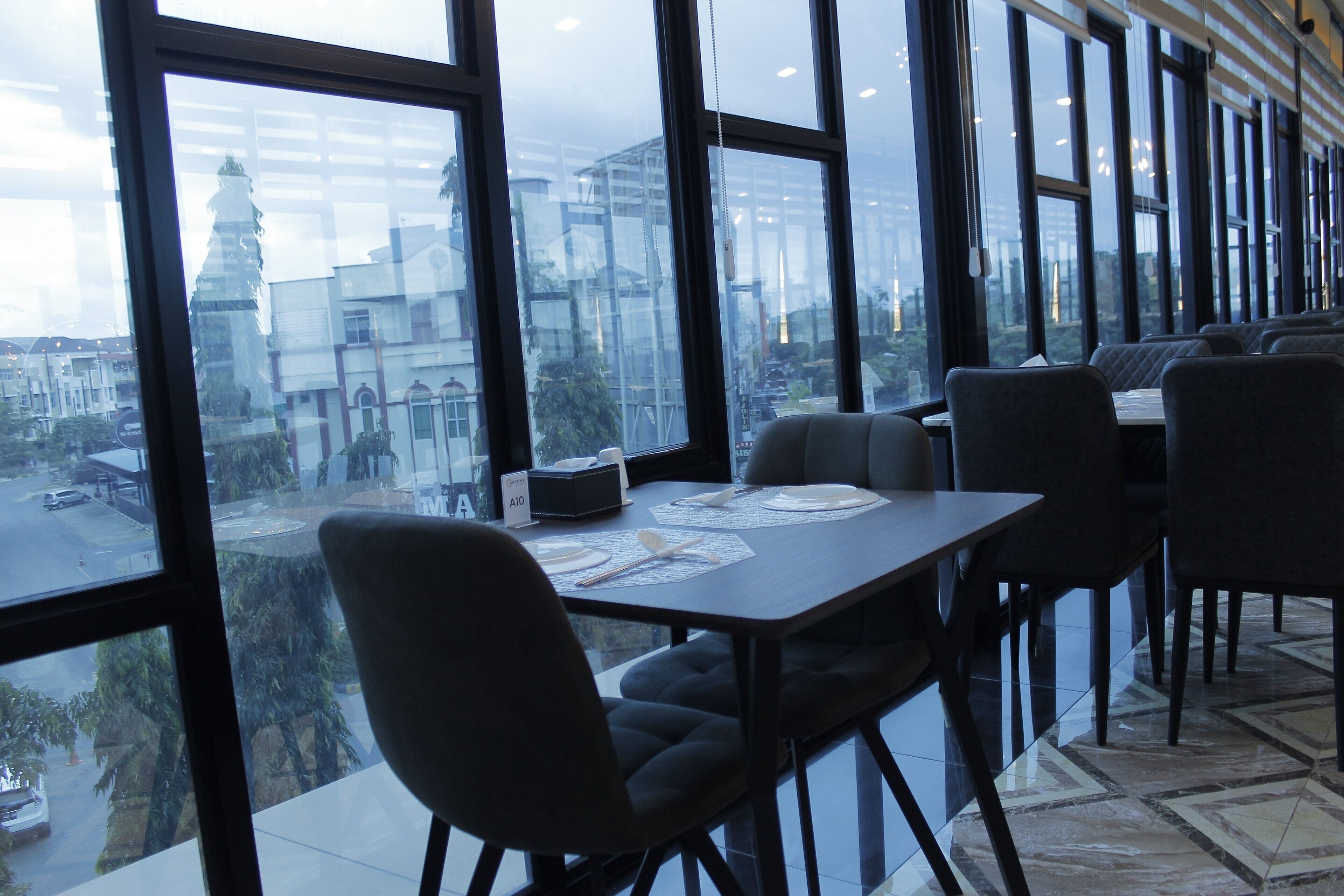 A restaurant with large windows, tables set with plates and cutlery, and a view of a cityscape.