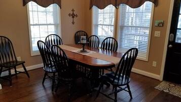 A dining room with a wooden table and six chairs, large windows with blinds, and a decorative wall piece.