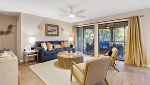 A modern living room with a sofa, coffee table, and chairs, a ceiling fan, and a view through large glass doors to an outdoor area.