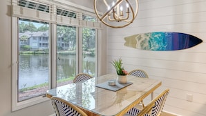 A dining area with a glass table, chairs with blue and white patterns, a chandelier, and a wall-mounted surfboard.