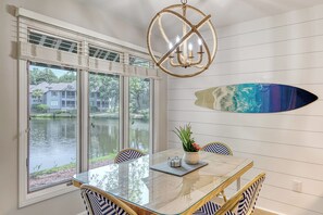 A dining area with a glass table, chairs with blue and white patterns, a chandelier, and a wall-mounted surfboard.
