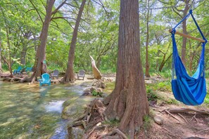 A serene forest setting with a clear stream, blue chairs, and a blue hammock.
