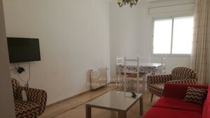 Una sala de estar con un sofá rojo, sillas estampadas, una mesa de centro de vidrio, una mesa de comedor con sillas, un televisor montado en la pared y un radiador.