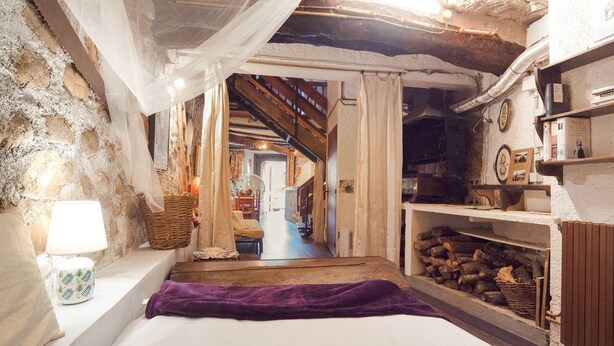 Una habitació rústica amb un llit, una làmpada, una llar de foc i una escala que porta a una altra habitació.