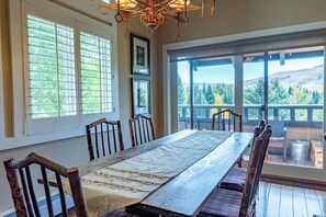 A dining area with a long table, wooden chairs, and large windows offering a view of trees and hills.