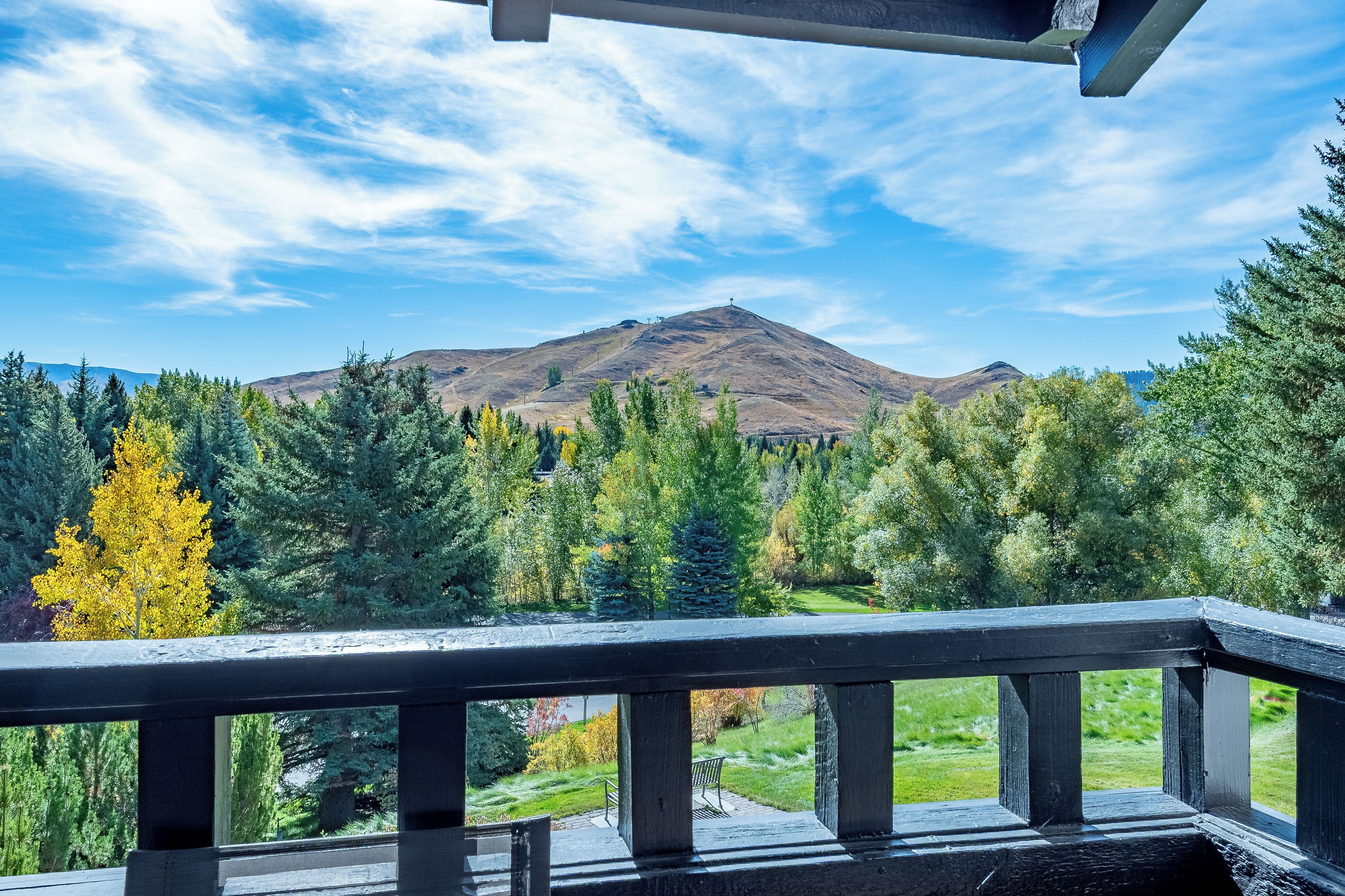 A mountain view from a balcony with a railing, overlooking a forested area and a hill with a structure on top.