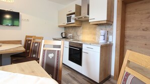 Cocina compacta con gabinetes blancos, microondas, cafetera y estufa. Hay una mesa de comedor de madera con sillas y un televisor montado en la pared.