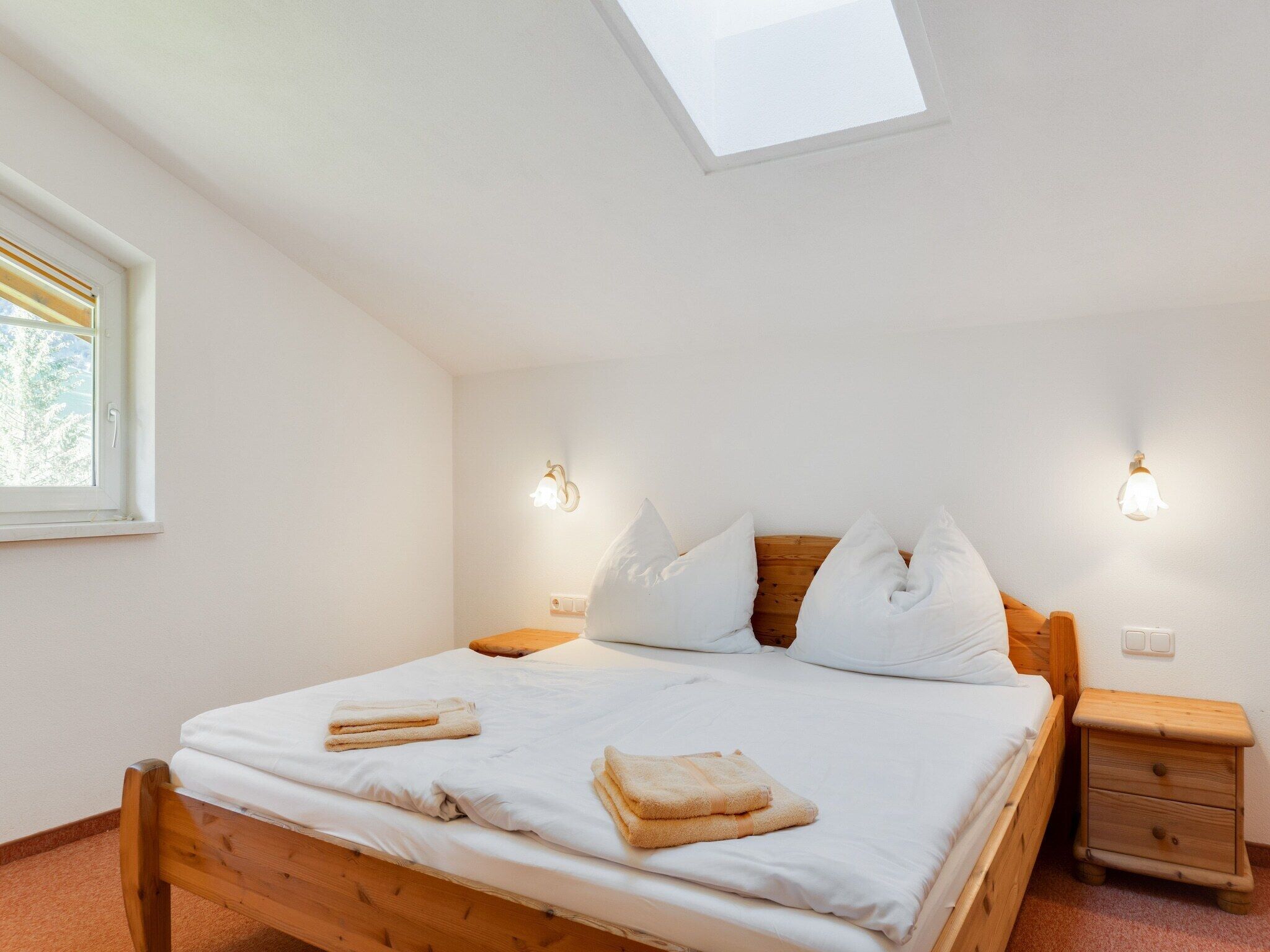 A bedroom with a wooden bed, white bedding, two bedside lamps, a nightstand, and a skylight.