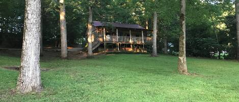 A wooden cabin with a deck, surrounded by trees and grassy area.